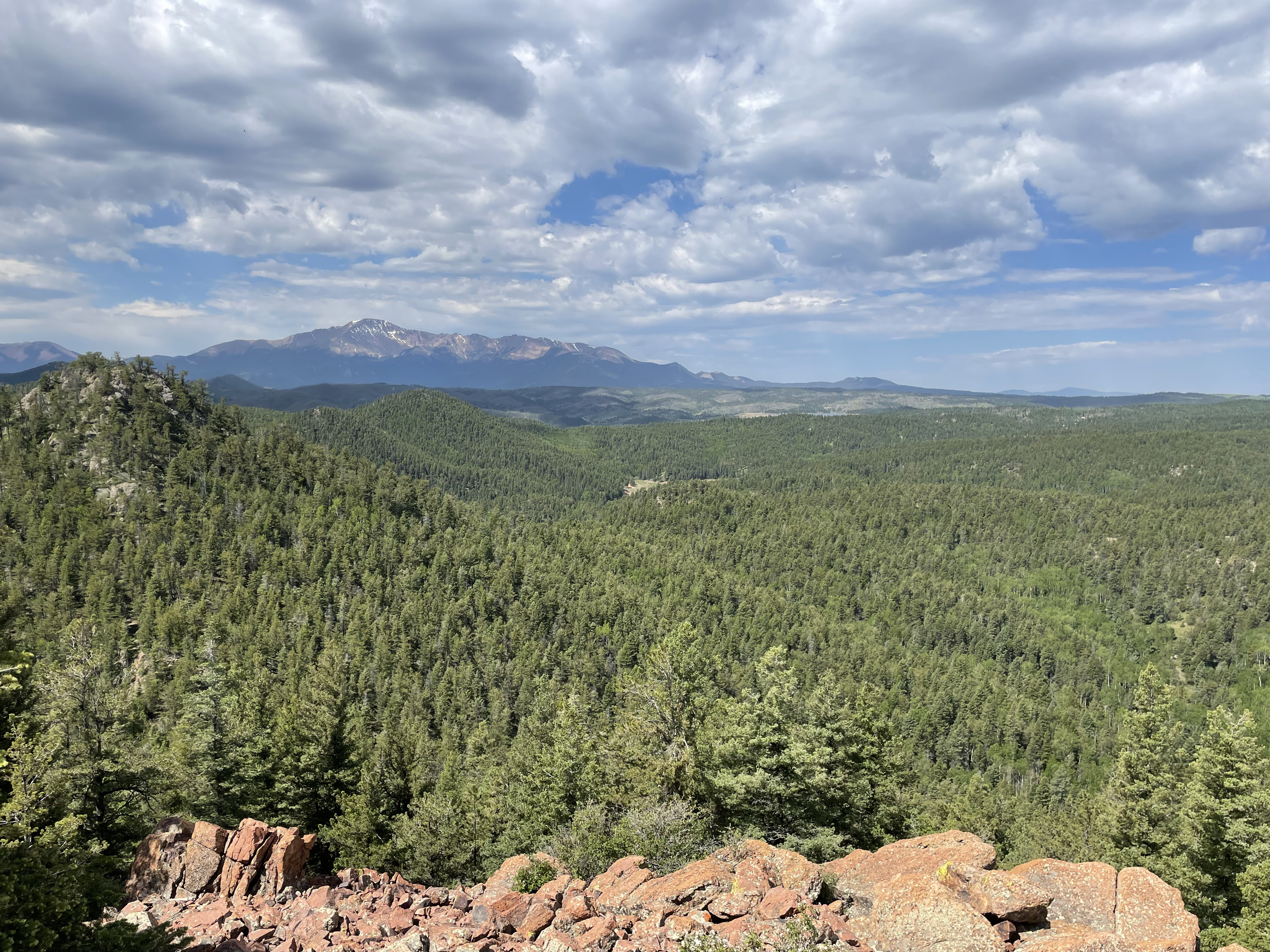 Scenic view from Eagles Peak in Colorado Springs. A vast expanse of dense green forest stretches across rolling hills. In the background, Pikes Peak stands with its partially snow-capped summit. The sky is a mix of blue and white with scattered clouds casting soft shadows over the landscape. The foreground features reddish-brown rocky terrain.